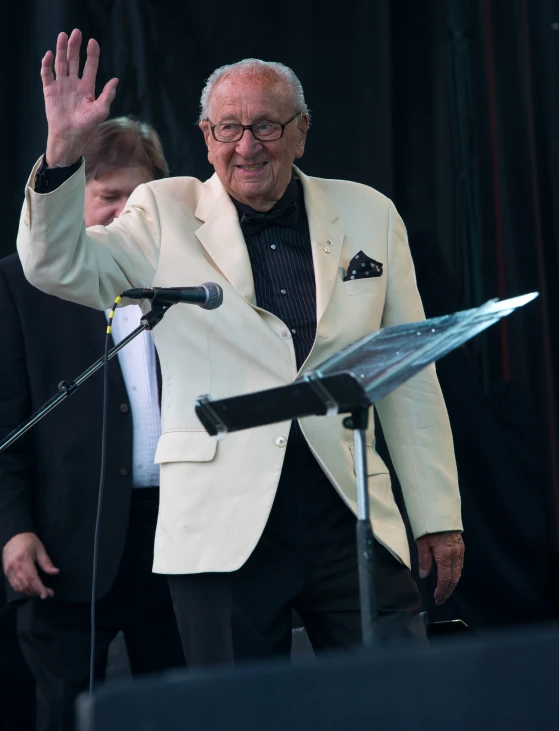 an older man waves at people as he stands behind him in front of microphones