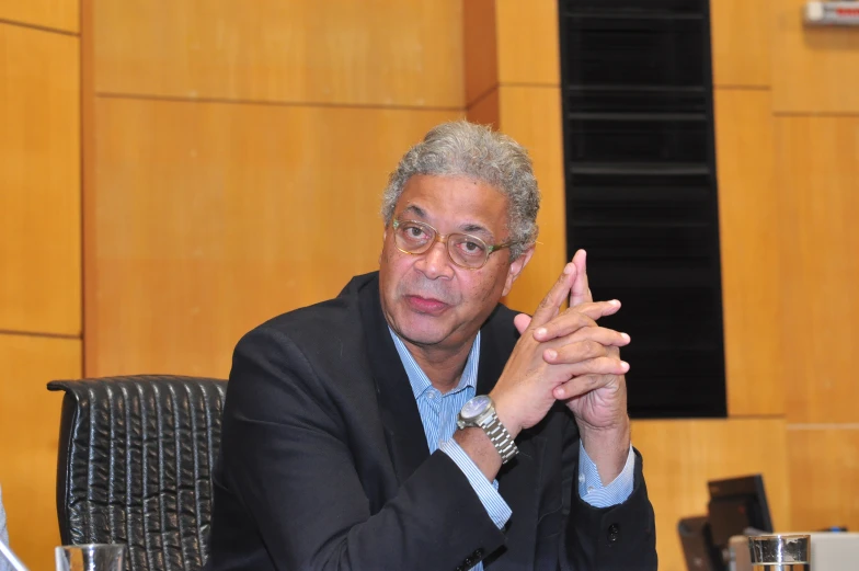 a man in a suit sits at a table with one hand raised up