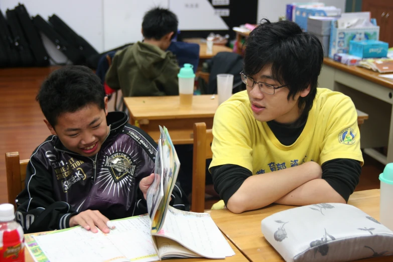 two boys sitting at a desk looking at an open book