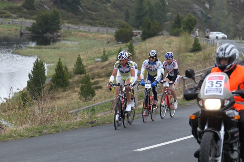 three cyclists with helmets, one on the front of the bike, are racing while another is standing beside a lake