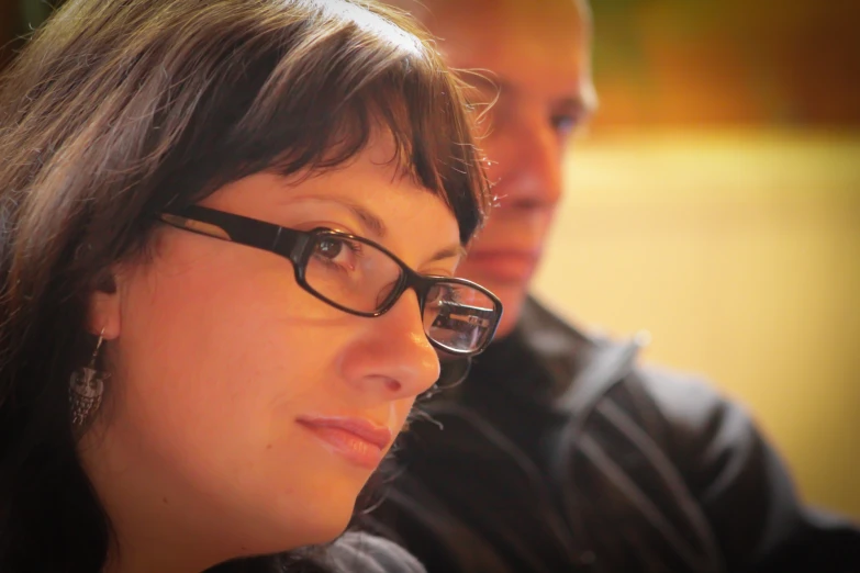 a woman with glasses sits at a table while another mans looks on