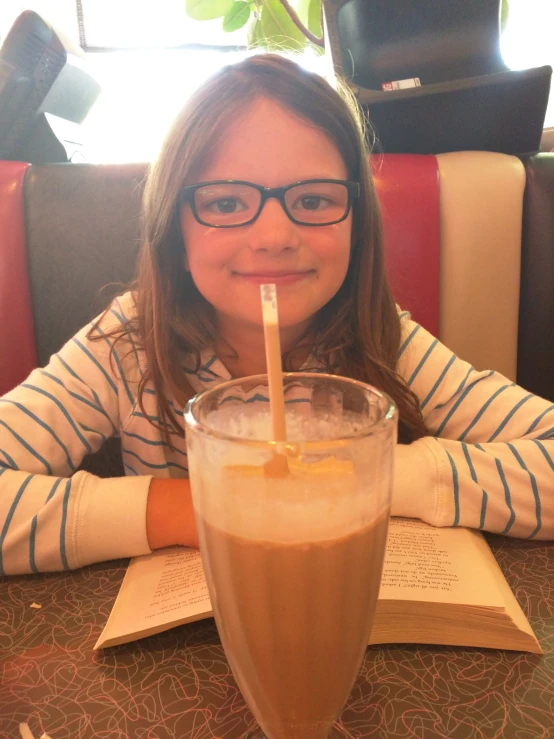 a girl wearing glasses sits at a table and drinks from a glass