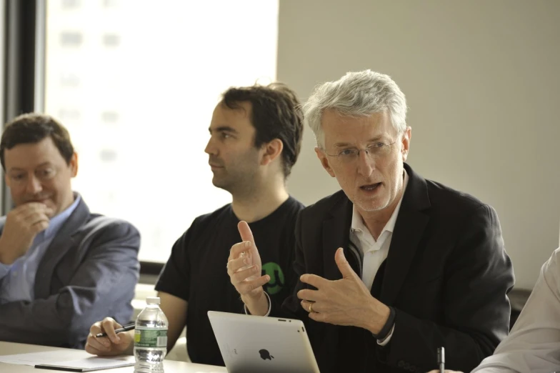 three men are sitting at a table and one of them is on his laptop