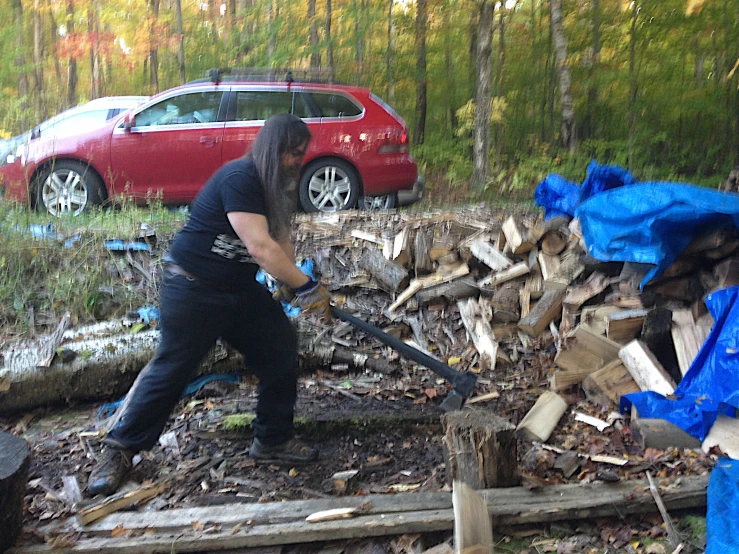 a man in a black shirt and blue pants holding an iron ax next to a pile of wood