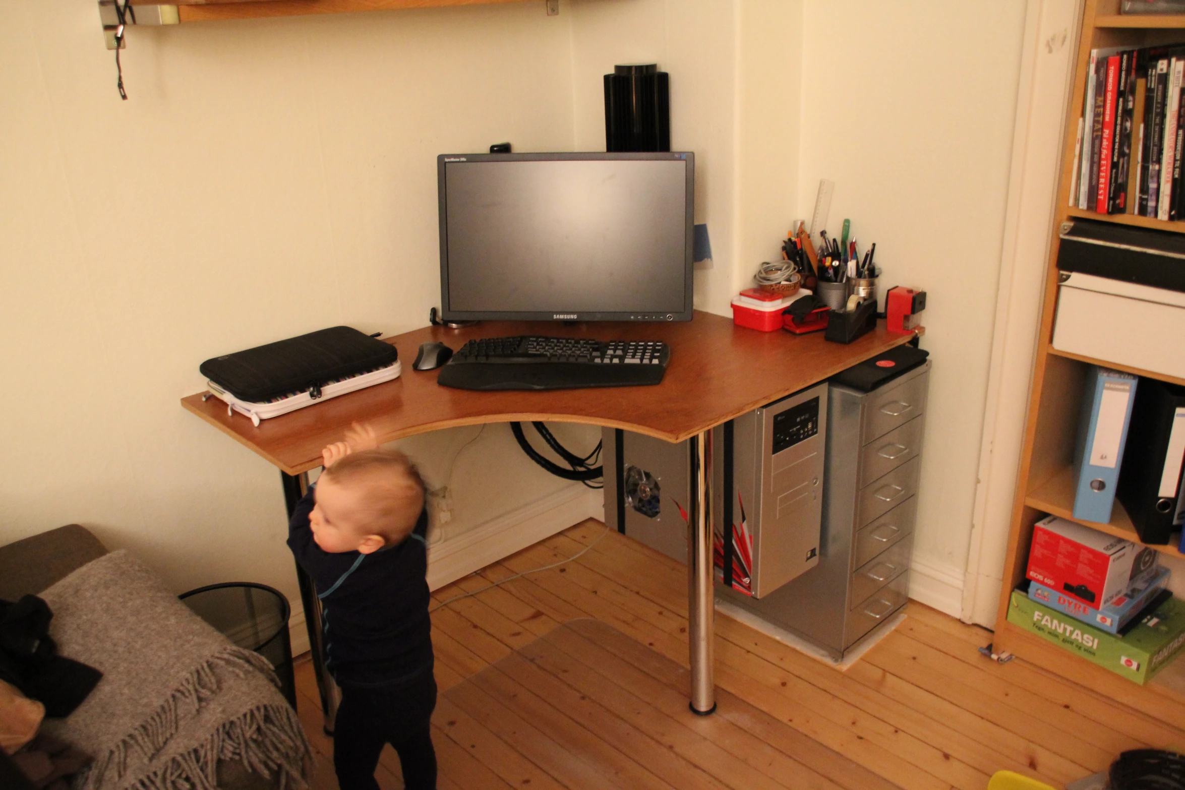 a little  standing at a computer on top of a wooden desk
