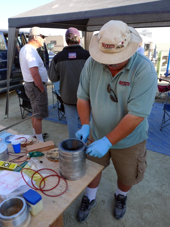 an older man in a hat and blue shirt works on an object on a table under a tent