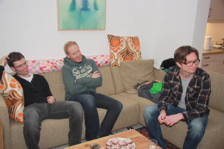 two men sit on sofas, one of them holding his hands up