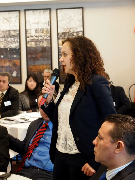 a woman standing in front of a microphone with other people seated