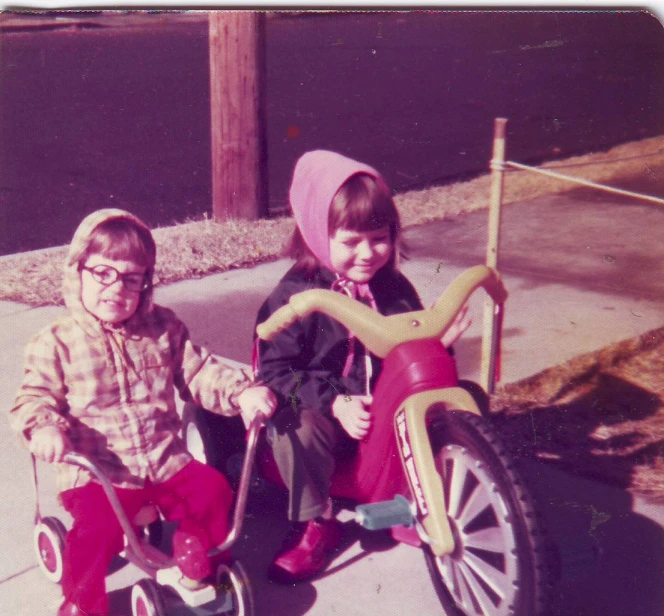 two children riding a red tricycle down the sidewalk