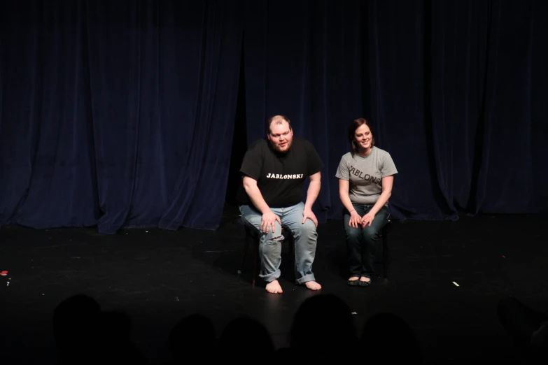 two people sit on a stage in front of a dark curtain