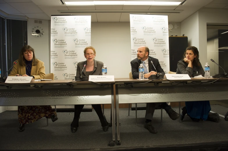 three people sit at a conference table, one person stands at the front