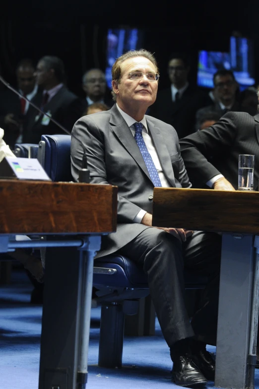 two men in suits sitting at tables in front of other people