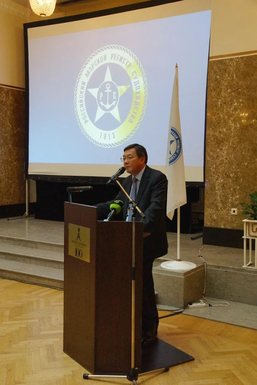 a man standing at a podium speaking in front of a projector