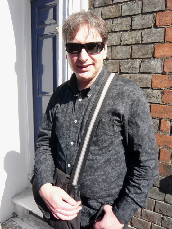 a man in sunglasses and grey sweater standing by brick wall