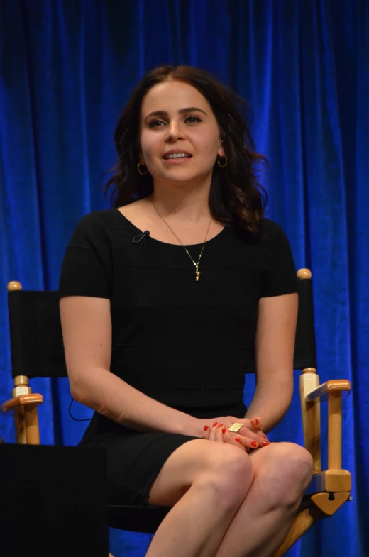 a woman in black sitting in a chair in front of a blue curtain