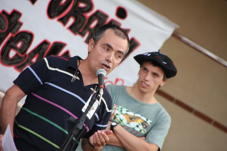 a man speaking into a microphone next to a male wearing a baseball cap