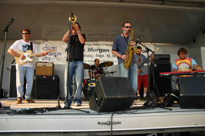 three men stand on stage as one plays the saxophone