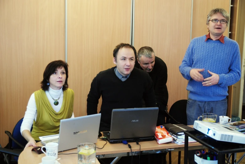 five people gathered around the desk while one man is typing on the computer