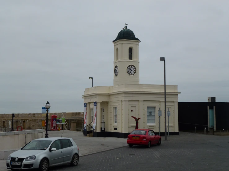 a clock tower stands over a small white building with a car parked in front of it