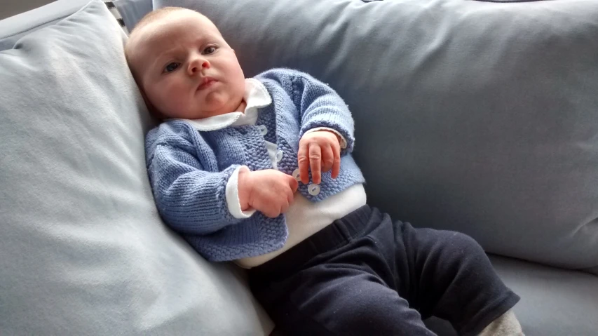 baby wearing blue sweater and dark pants on gray couch
