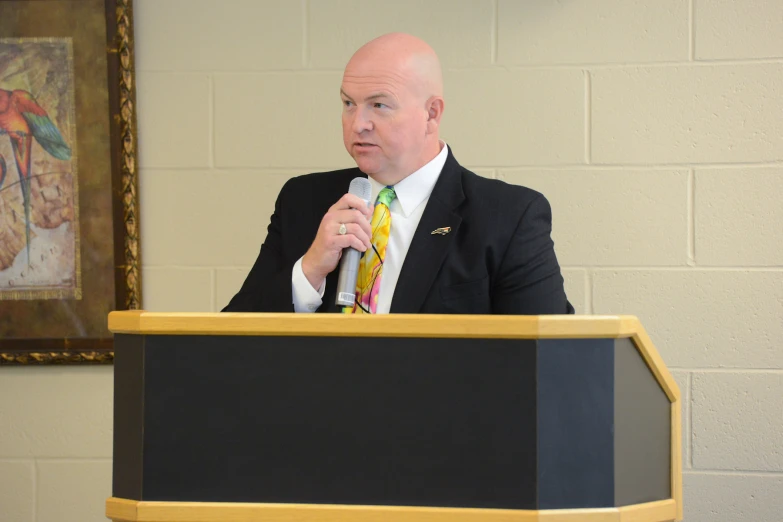 a bald man in a suit stands behind a podium while holding a microphone