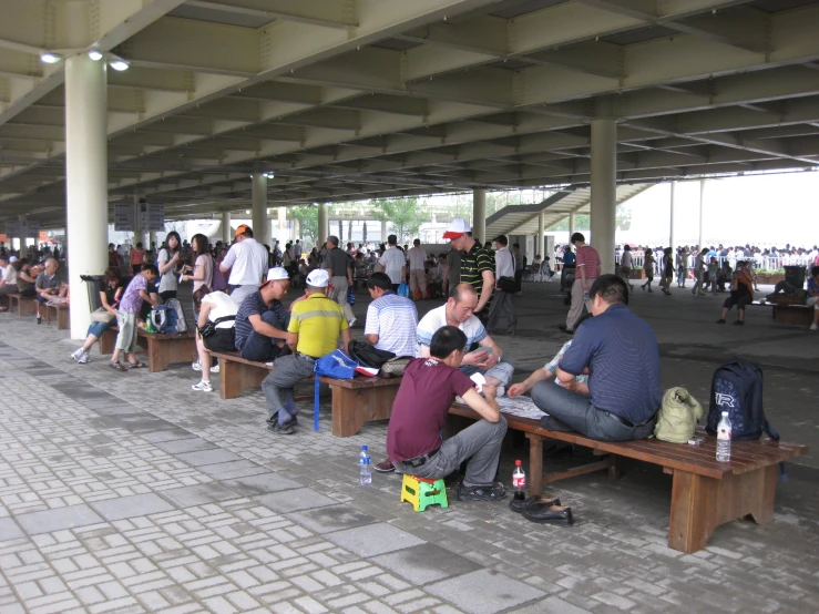 a crowd of people sitting around benches under a large metal roof