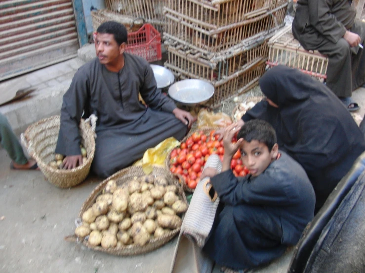 three men sit on the ground next to baskets of potatoes and tomatoes