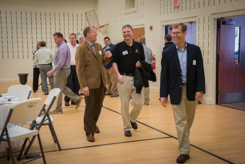 three men walk inside a gym with tables and chairs on the floor