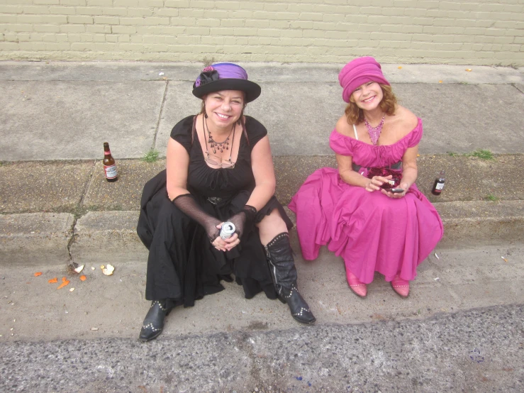 two women sit on the sidewalk wearing long dress with hats and boots