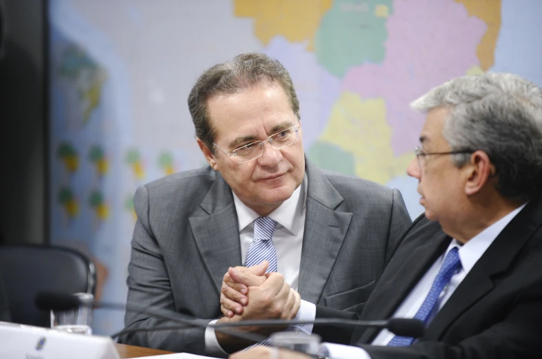 a man sitting at a desk in front of another man who is holding his hand together