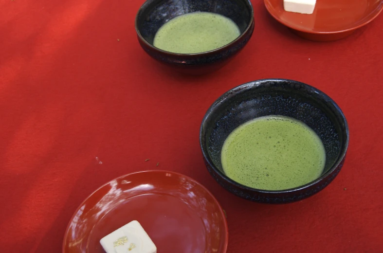 two black bowls with green soup next to a plate with a piece of cheese