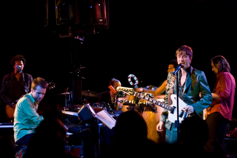 man standing with guitar, singing into microphone while other men sit