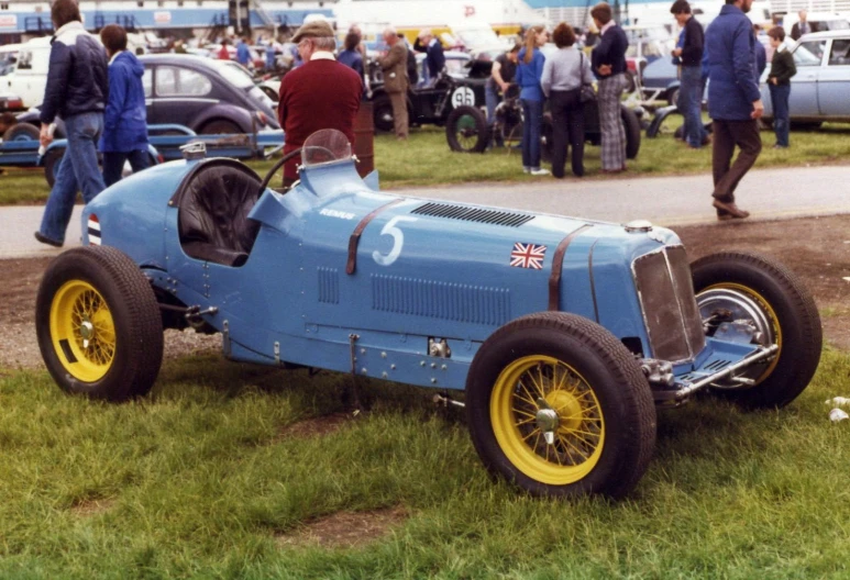 a large blue race car with yellow tires and yellow wheels is parked on grass while other cars are in the background