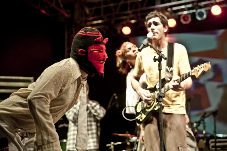 a man in a red mask and a band plays with guitar