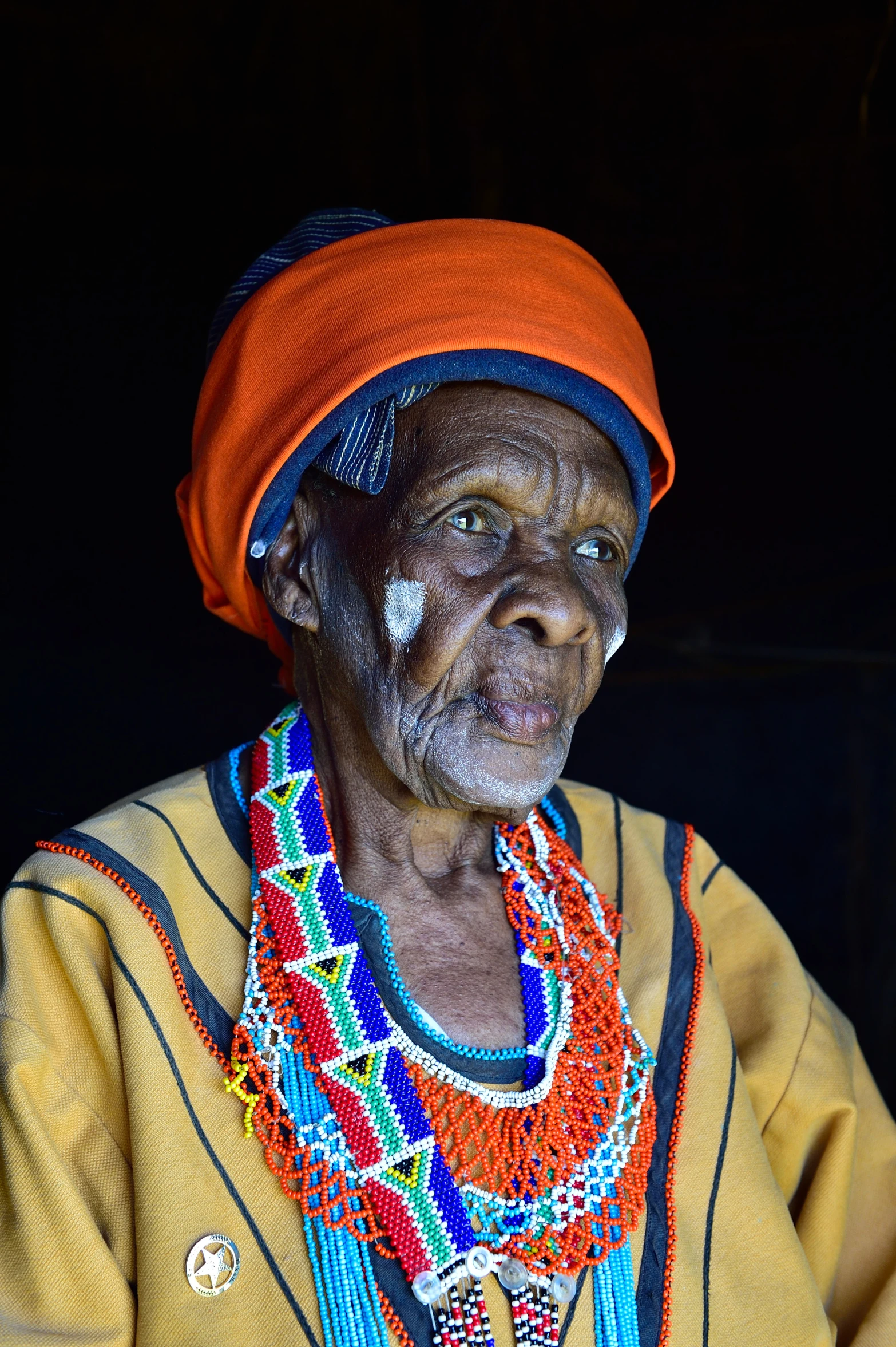 an elderly woman in brightly colored necklaces sits with dark background