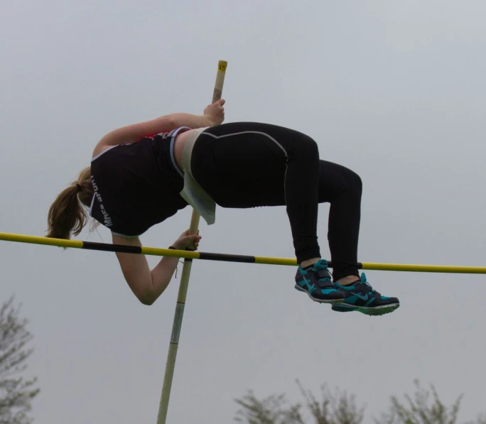a female high jumper balances on the bar with trees in the background