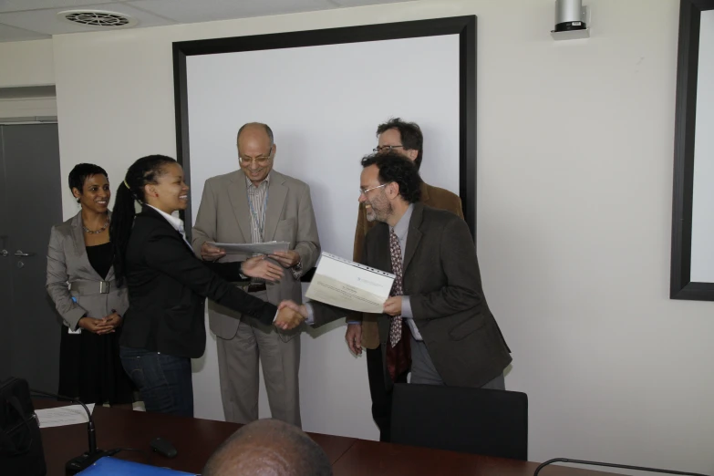 four people standing in a room, two shaking hands, one person in suit and tie