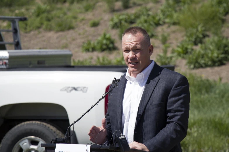 a man speaking from a podium outside with a truck in the background