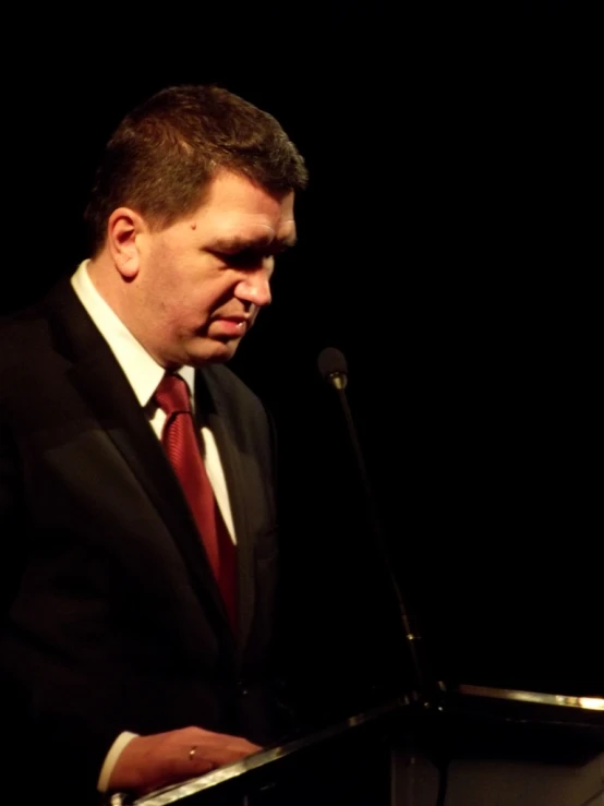 man in dark suit and red tie standing behind a podium