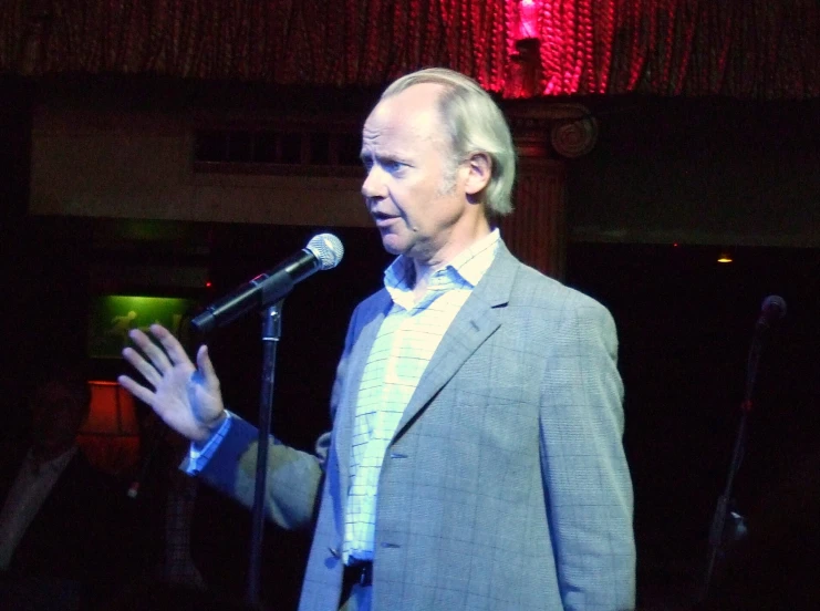 the man stands at a microphone and holds his hands out while holding a drink