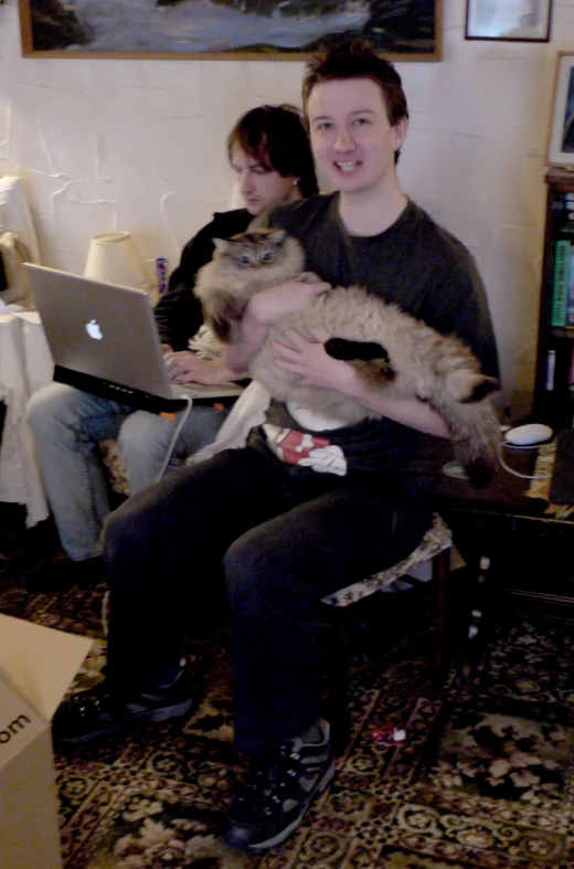 two people sit on a couch in front of a laptop, and holding a cat