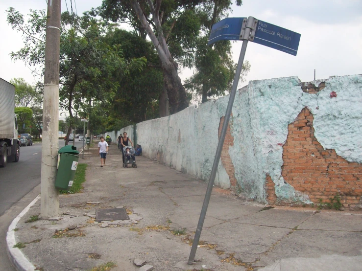 people walking on a side walk past an old wall