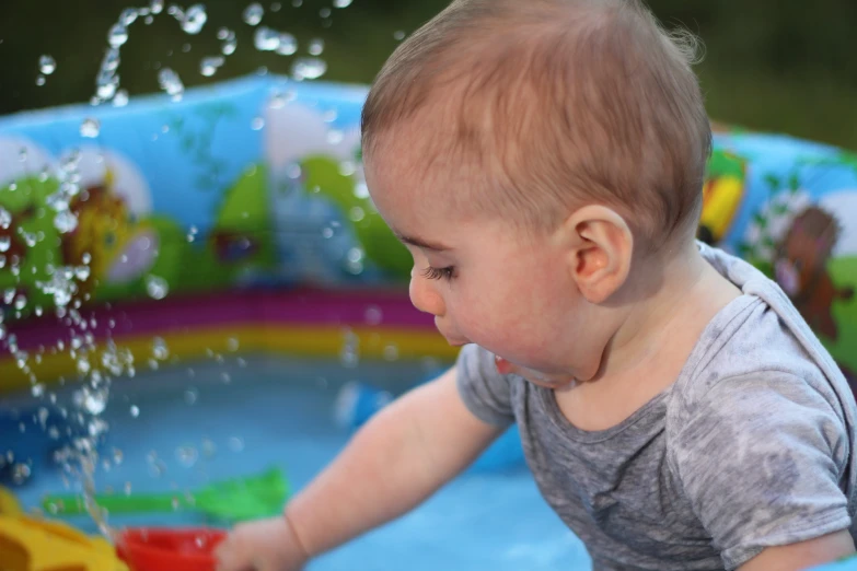 a young child in grey shirt splashing water on a colorful baby in blue and yellow pool