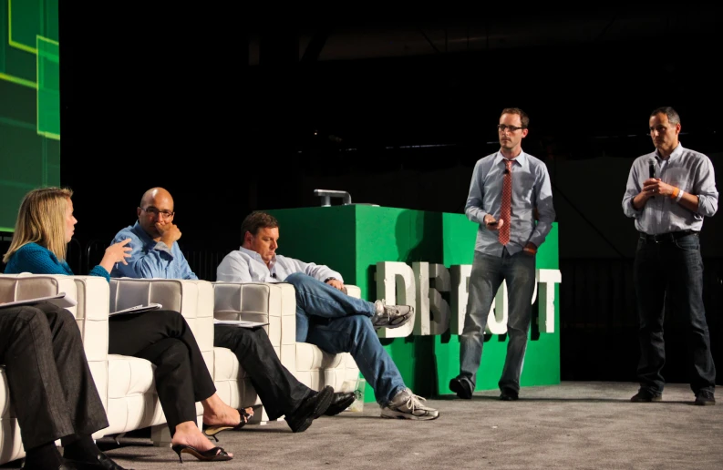 three men and one woman talking on stage with people seated