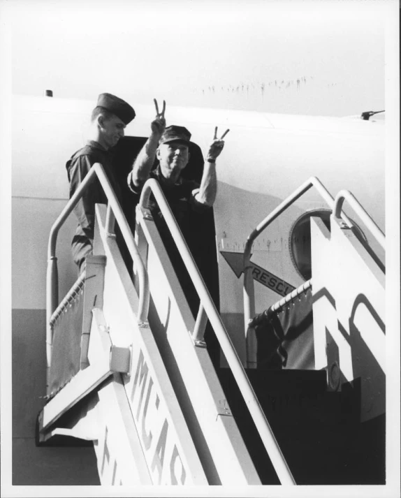 two men are standing on an airplane deck, giving a sign