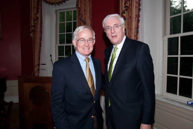 two men wearing suits and tie standing by a window