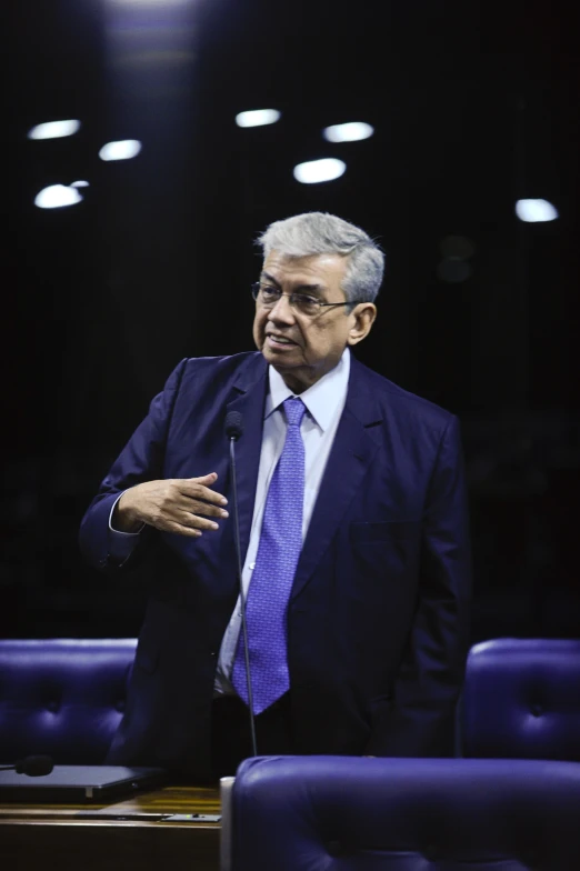 a man stands at the microphone wearing glasses and a suit