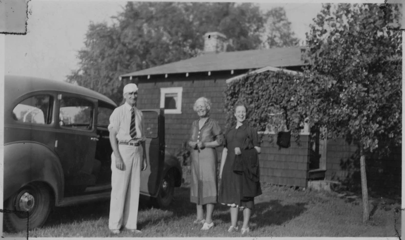 two women and an old man are standing in front of a house