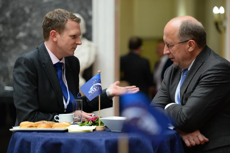 two men sit at a table talking and eating