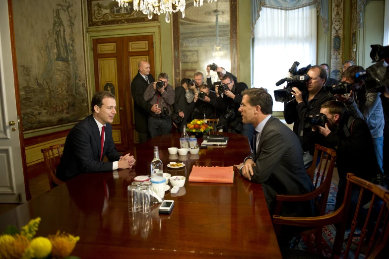 two men sit at a wooden table, with cameras on their backs